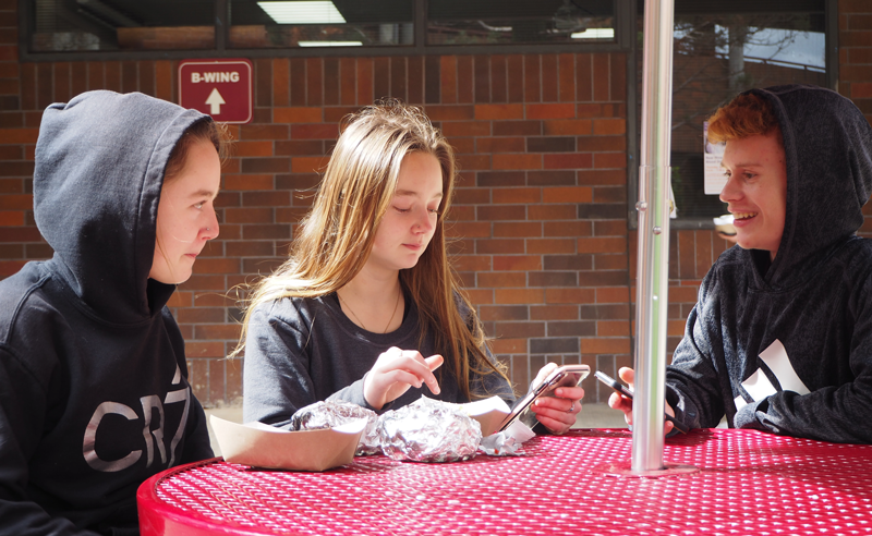 Freshmen Jade and Mariah Thibodeau, along with fellow Freshmen Sam Evans, put up their hoods to keep out the rain as they sit at a covered table.  Photo by Maddie Meilinger. 